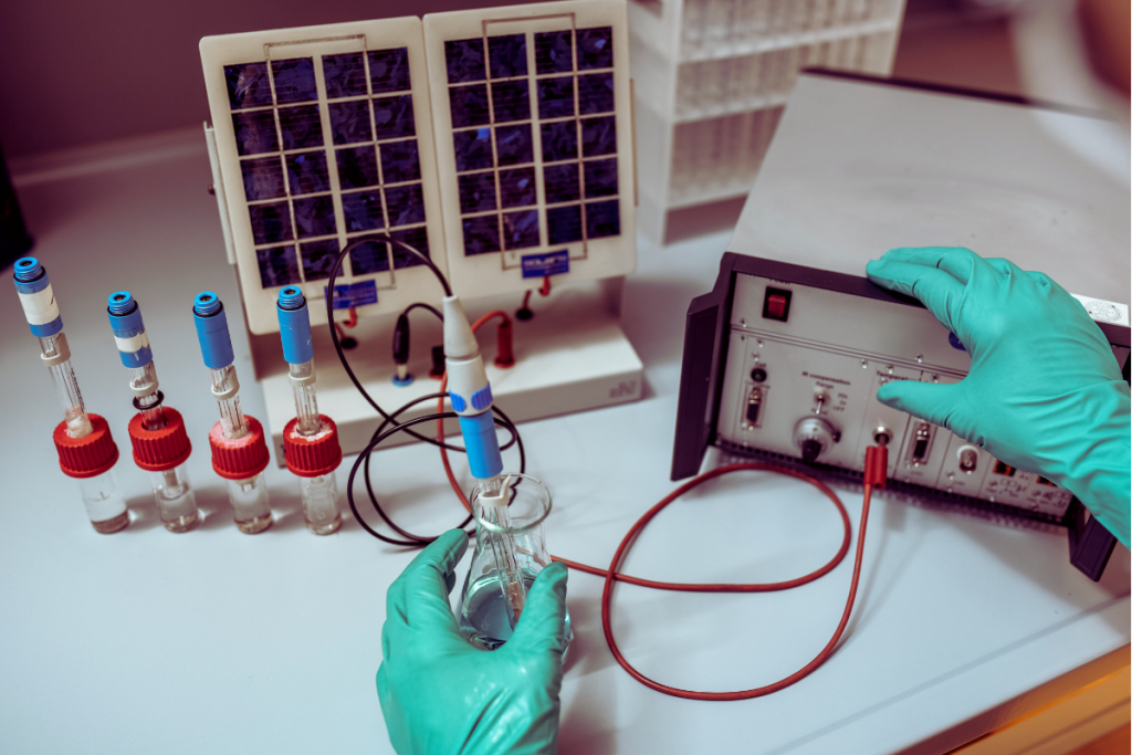 Stock image of gloved hands in a chemistry lab performing an electrochemistry experiment with a voltmeter, flasks, and tubes.